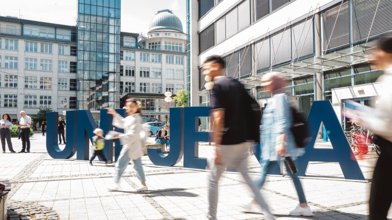 Platzhalterbild — Personen laufen vor den Uni Jena Buchstaben über den Campus